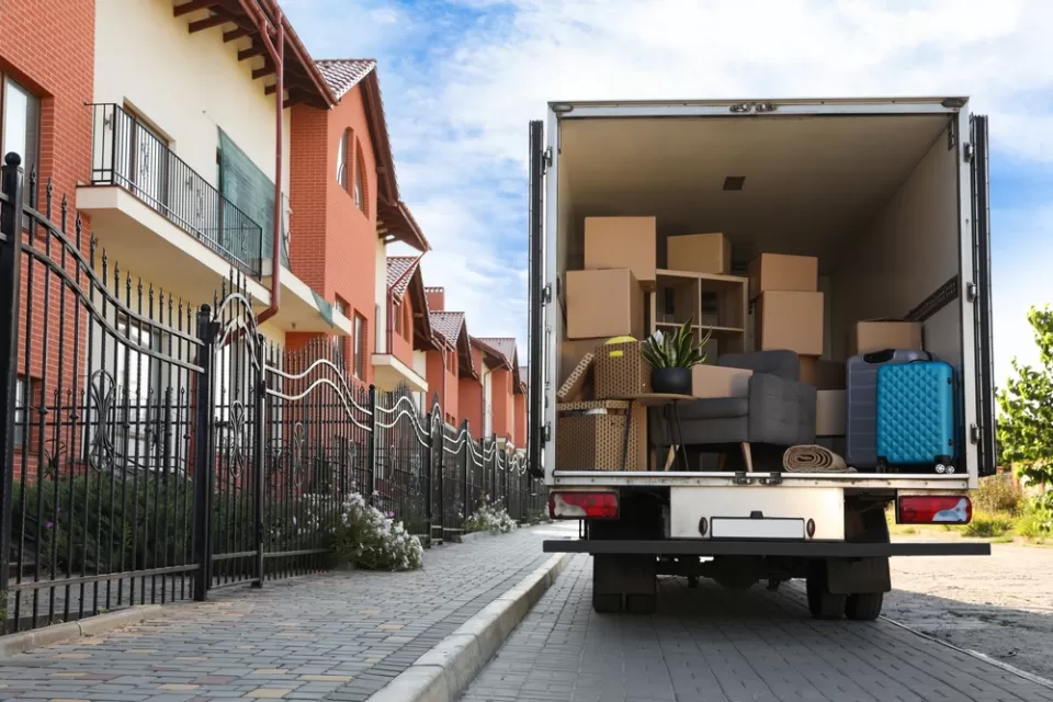 View of back of moving van with back door open, loaded with boxes, next to a line of two-story apartment buildings.